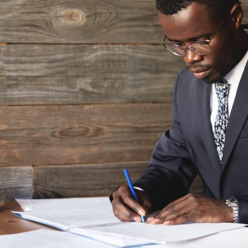 Serious black corporate worker in formal suit and spectacles signing a lucrative contract with concentrated expression while having morning cappuccino at a cafe, sitting against wooden wall background